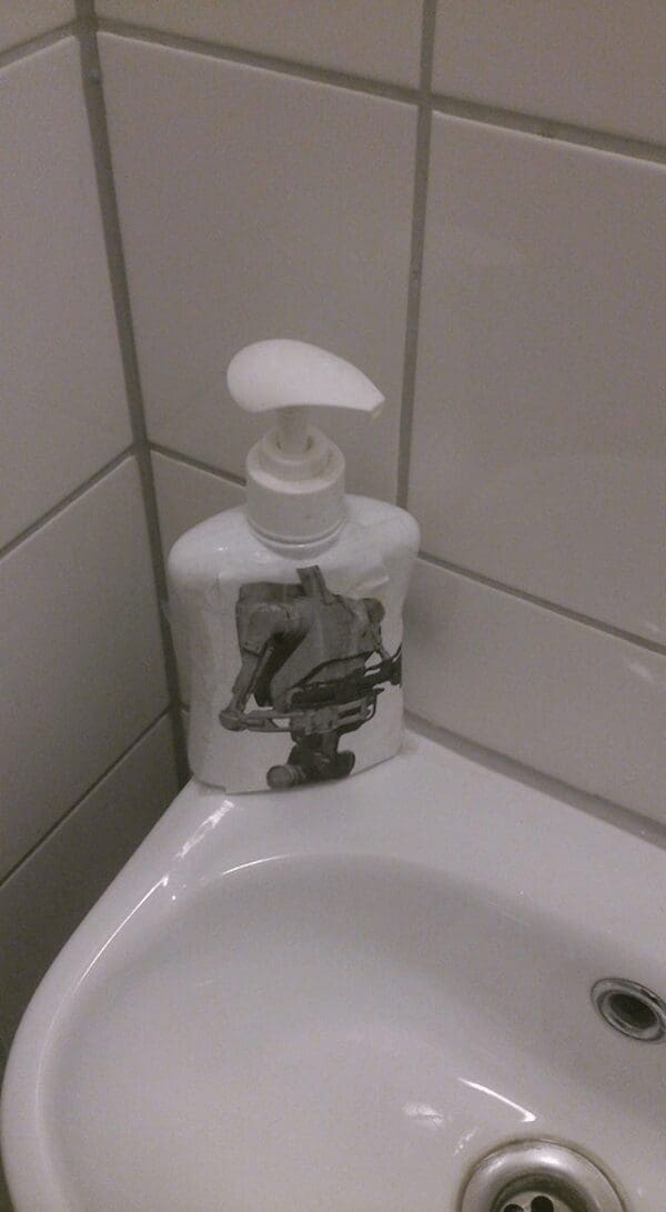 real-life star wars meme showing a printed picture of a B1 Battle Droid taped perfectly onto a white bathroom soap dispenser, making the pump look exactly like the droid's long head.