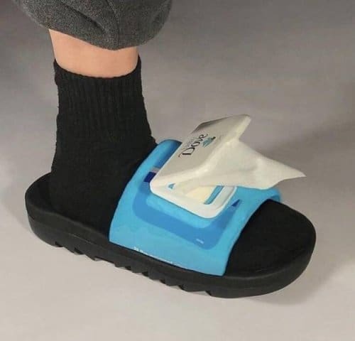 A "cursed" piece of footwear where the strap of a black slide sandal has been replaced by a functional pack of Dove wet wipes, complete with a plastic flip-top lid and a protruding tissue.