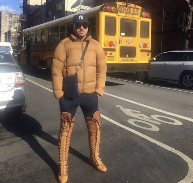 A city dweller stands on a sidewalk in a tan puffer jacket and black joggers paired with experimental thigh-high, lace-up boots that mimic the design of classic tan Timberland work boots.