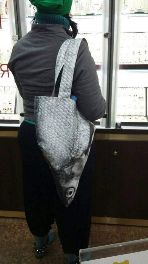 A shopper carries a tote bag featuring a hyper-realistic, oversized print of a fish head, making it look as though a giant, scaly sea creature is hanging vertically from their shoulder.