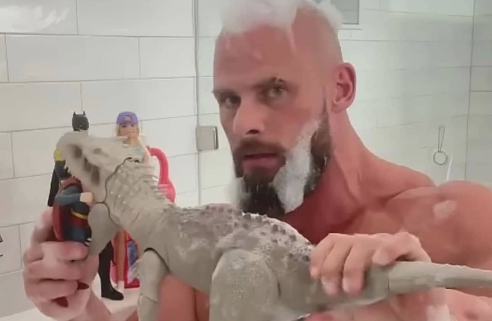 Muscular man with soap foam beard holding Batman and Superman figures near a toy dinosaur.