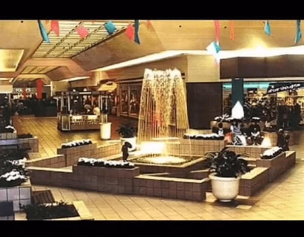 A beautiful 90s mall glamour shots centerpiece featuring a tiered brick fountain. Water cascades in thin, lit-up streams, surrounded by built-in brick planters and large white pots. Colorful pennant banners hang from the ceiling, adding a festive atmosphere to the wide, open walkway.