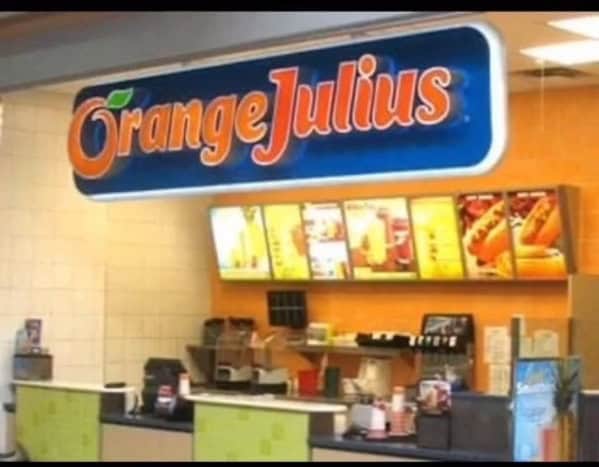 A vibrant 90s mall glamour shots photo of an Orange Julius kiosk in the food court. The glowing blue and orange sign dominates the frame, overlooking a bright yellow menu filled with classic fruit smoothies and hot dogs.