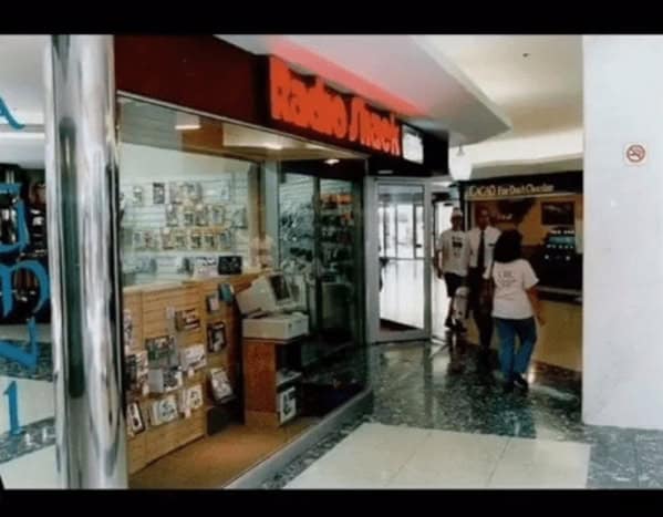 technical 90s mall glamour shots look at the entrance to a Radio Shack. The window display features bulky beige CRT monitors and desktop towers, set against a backdrop of the era's signature green marble-patterned mall flooring.