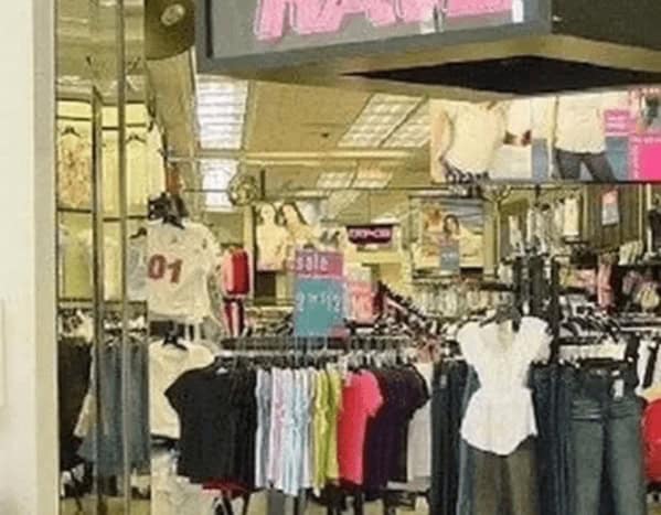 stylish 90s mall glamour shots entry showing the interior of a trendy 90s clothing store. Racks are packed with $12 graphic tees and brightly colored apparel, reflected in mirrored walls under the harsh, theatrical glow of mall spotlights.