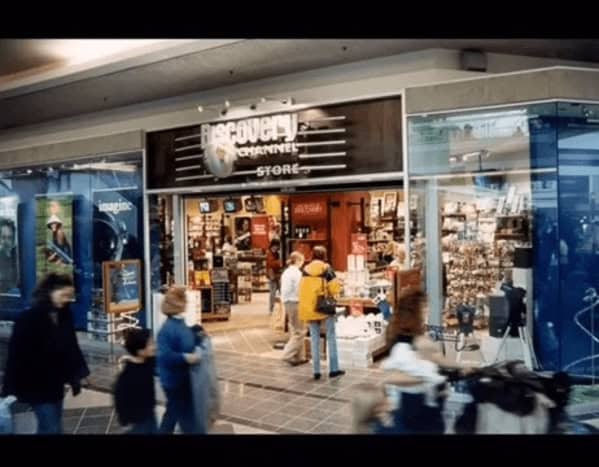 A sleek 90s mall glamour shots photo of The Discovery Channel Store. The storefront features a metallic logo and window displays of telescopes and space-themed posters, attracting curious shoppers into its high-tech, educational interior.