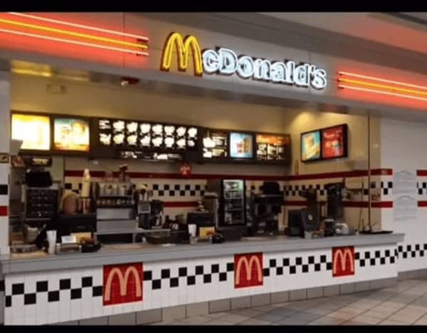 A classic 90s mall glamour shots counter view of a food-court McDonald's. The design features the iconic black-and-white checkered tile and a warm yellow neon "Golden Arches" glow over the registers and menu boards.
