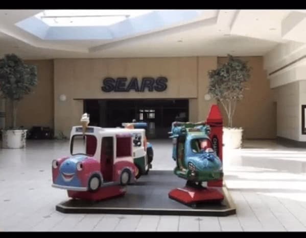 A moody and slightly liminal 90s mall glamour shots capture of a Sears department store entrance. In the foreground, colorful coin-operated kiddie rides—including a pink bus and a blue helicopter—stand idle on a carpeted platform under the soft glow of a skylight.