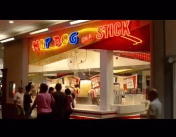 A high-energy 90s mall glamour shots capture of a Hot Dog on a Stick. The bright red and yellow neon sign glows above a counter crowded with shoppers waiting for fresh lemonade and corn dogs.