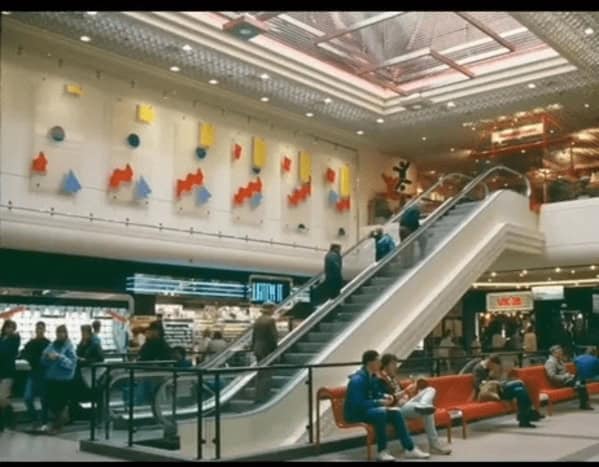 This 90s mall glamour shots entry highlights the grand architecture of a multi-level shopping center. A long escalator carries shoppers past abstract, colorful geometric wall art. In the foreground, people rest on iconic curved orange benches, surrounded by the warm, filtered light of a massive atrium.