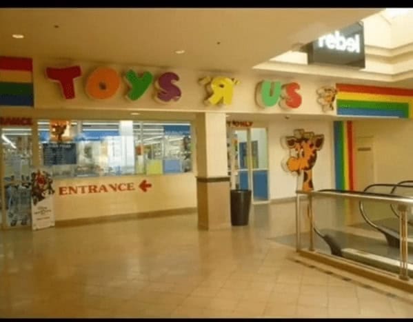 classic 90s mall glamour shots storefront for Toys "R" Us. The entrance features the iconic rainbow-striped branding and a large cutout of Geoffrey the Giraffe. The polished tan tile floors and bright fluorescent lighting evoke the excitement of a childhood shopping trip.