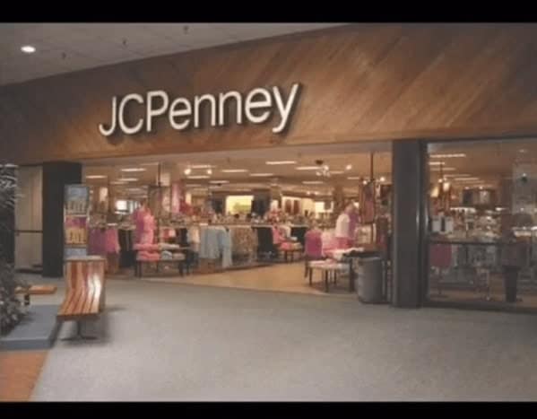 quintessential 90s mall glamour shots photo of a JCPenney entrance. The storefront is defined by heavy dark wood paneling and the classic sans-serif white logo. Inside, racks of brightly colored 90s apparel are visible, creating a warm, inviting glow against the cool grey floor.