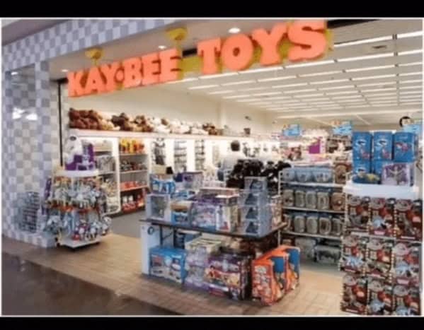 A vibrant 90s mall glamour shots look at a Kay-Bee Toys store. The bright orange-and-yellow bubble-letter sign hangs over shelves packed with classic toys, action figures, and a massive wall of plush stuffed animals. The signature checkered flooring adds to the frantic, playful energy.