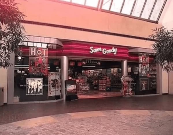 peak 90s mall glamour shots view of a Sam Goody music store. The entrance is framed by bright red neon stripes and posters of boy bands and grunge icons. The dark interior and polished stone floors represent the ultimate teenage hangout spot of the decade.