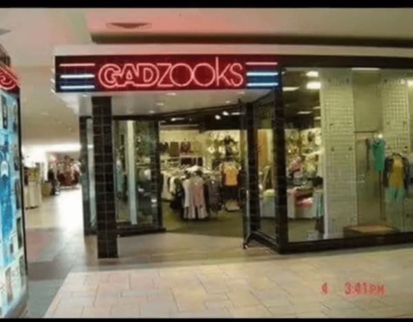 90s mall glamour shots capture of a Gadzooks storefront. The name glows in red and blue neon, reflecting off glass displays filled with oversized jeans, graphic tees, and alternative 90s fashion. The interior is a dense, high-energy mix of industrial lighting and pop culture merchandise.