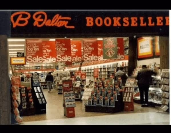 A busy 90s mall glamour shots perspective of a B. Dalton Bookseller. The store is plastered with red "Sale" banners, and the shelves are packed from floor to ceiling. Shoppers in dark coats browse the narrow aisles under the glow of the iconic orange cursive logo.