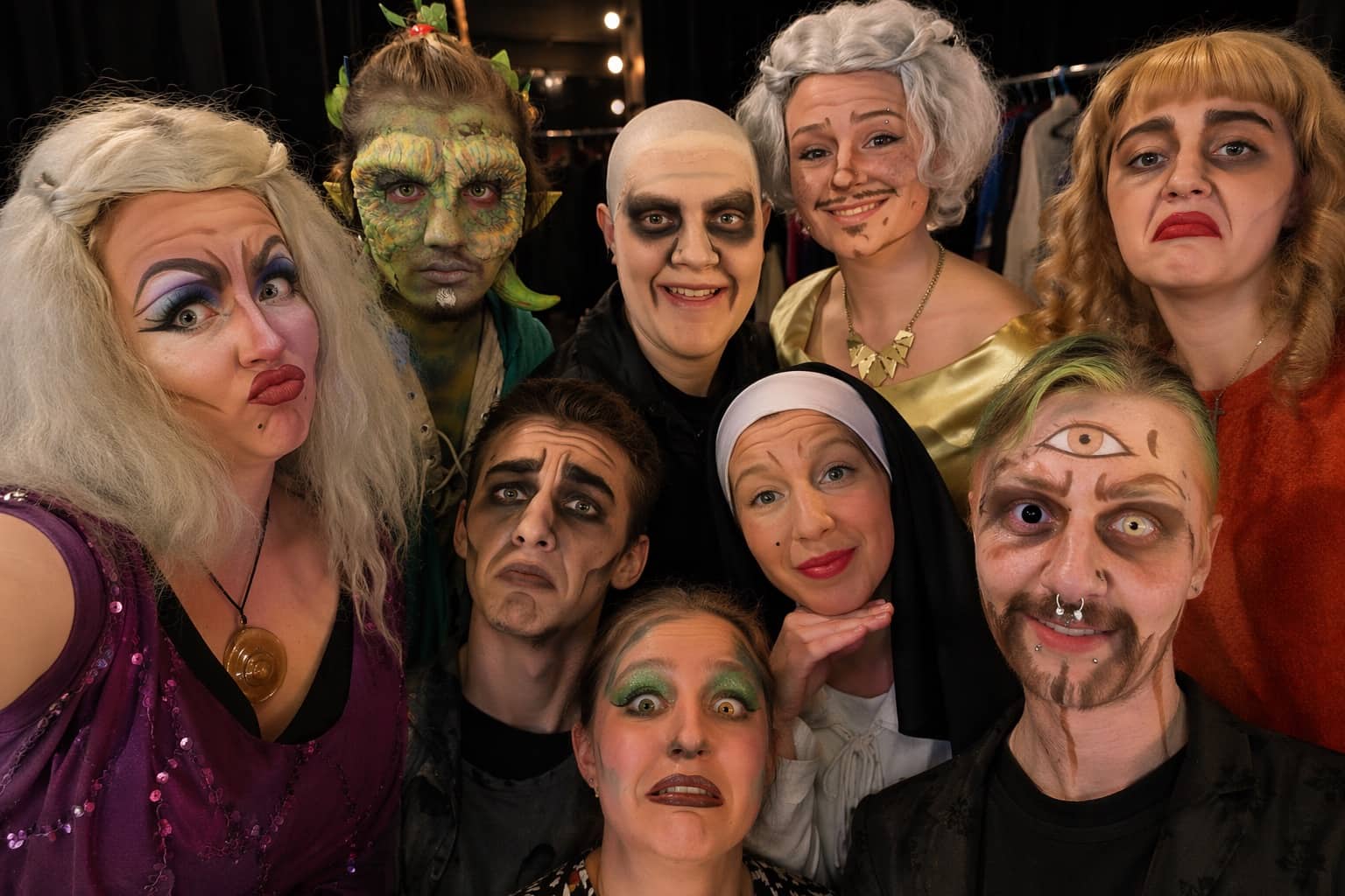 Theater troupe backstage wearing eccentric stage makeup and costumes including a green monster and nun.