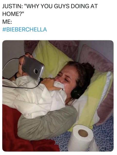 A Bieberchella meme featuring a person lying in bed, clutching a white comforter and crying while wearing headphones and staring intensely at a smartphone. The text above reads, "JUSTIN: 'WHY YOU GUYS DOING AT HOME?' ME: #BIEBERCHELLA," capturing the emotional intensity of fans watching the Coachella livestream from their rooms.