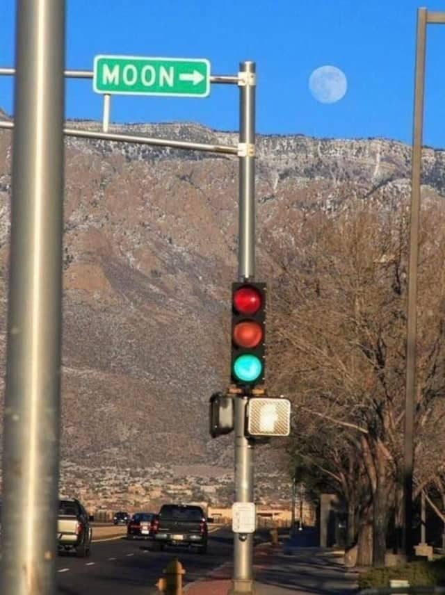 Photo showing a street sign pointing directly toward the moon visible behind it in daylight sky.