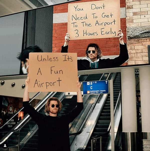 A street-style travel meme featuring a man holding up two cardboard signs in an airport. The first sign claims you don't need to arrive three hours early, while the second sign provides the crucial exception: "Unless It's A Fun Airport."