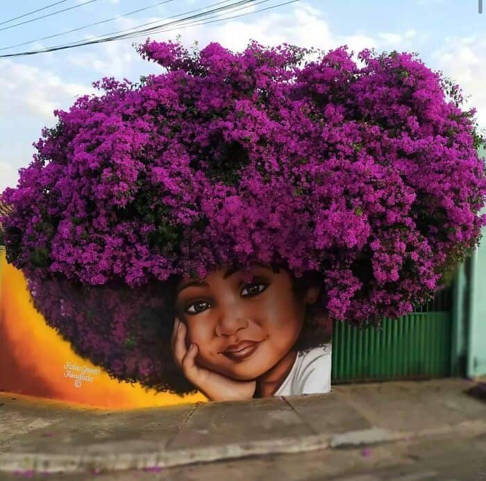 A stunning piece of amazing street art where nature meets paint. A mural of a young girl with a thoughtful expression is painted on a wall directly beneath a massive, vibrant purple bougainvillea bush. The blooming flowers perfectly mimic a lush, floral afro, creating a seamless 3D interaction between the artwork and the environment.