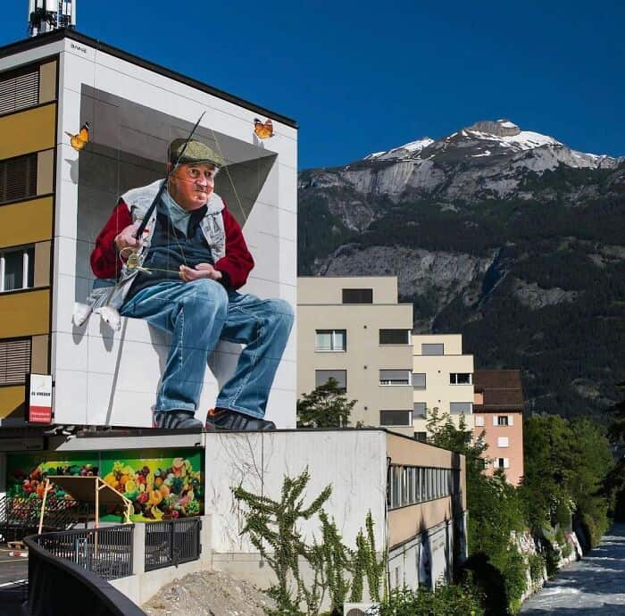 A mind-bending 3D mural of an elderly fisherman. He is painted sitting on a white ledge that appears to protrude from the side of a building, holding a long fishing rod. The background landscape features real mountains, making it look as though he is giant-sized and fishing directly into the valley below.