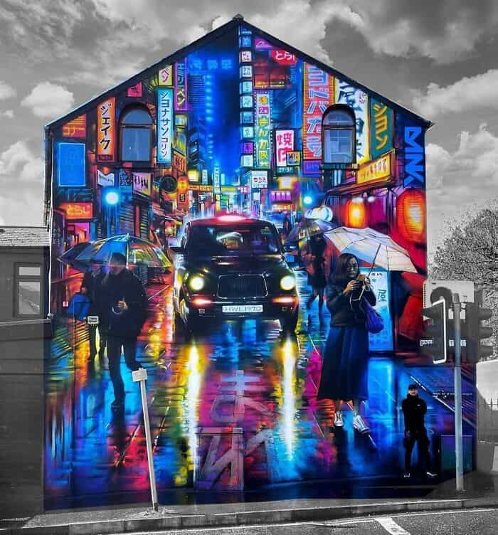 A breathtaking gable-end mural that transforms a quiet building into a vibrant, neon-lit Tokyo street at night. The amazing street art features a black taxi splashing through puddles, glowing signage in Japanese kanji, and pedestrians with umbrellas, all rendered with such depth that the wet pavement seems to shimmer against the real-world grey sky.