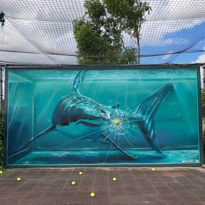 A clever 3D illusion painted on a wall behind a sports court. The amazing street art depicts a Great White shark in a massive tank; a painted tennis ball appears to have just struck the "glass," creating a realistic spiderweb of white cracks. Real tennis balls scattered on the pavement in the foreground blur the line between art and reality.