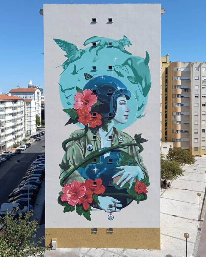 A towering vertical mural on a white apartment block featuring a woman in a vintage blue pilot’s helmet. She protectively cradles a glowing globe, which is being reclaimed by thick green vines and bright red hibiscus flowers. Above her, silhouettes of a fox, a bird, and a dolphin leap across a teal sky, symbolizing a harmonious fusion of humanity and nature.