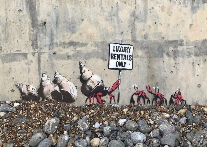 A poignant and satirical piece of amazing street art on a stained concrete wall above a bed of pebbles. A line of red hermit crabs is shown house-hunting; a larger crab points a claw toward a hand-painted sign that reads "LUXURY RENTALS ONLY," serving as a biting urban commentary on the global housing crisis.