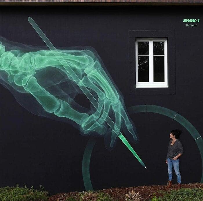 A striking "Radium" mural by artist SHOK-1. Against a deep black wall, a glowing, translucent green X-ray of a human hand is shown precisely gripping a stylus to draw a circle. The neon-green skeletal detail is so fine it looks like it’s emitting actual light, especially contrasted against a real person standing nearby.