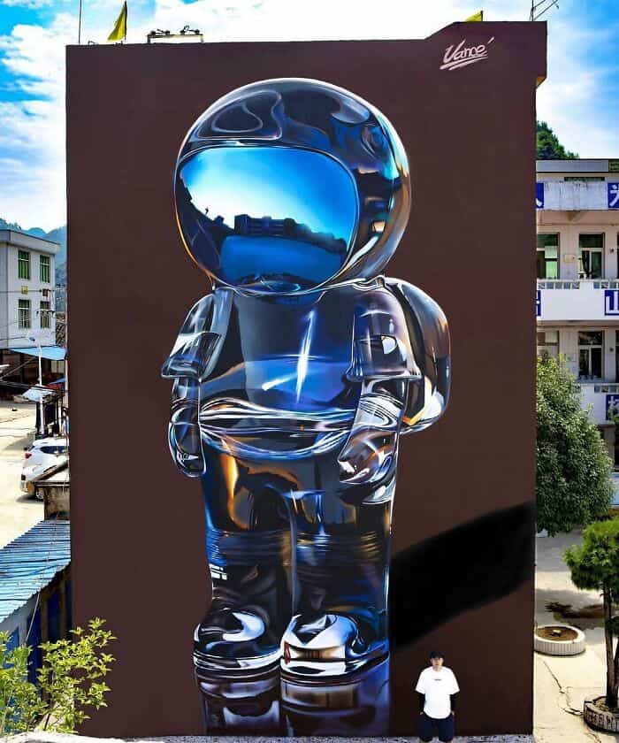 A masterclass in texture and reflection. This amazing street art depicts a small astronaut figurine on a chocolate-brown background. The suit is painted with a hyper-realistic "liquid chrome" finish, perfectly capturing the distorted blue sky and surrounding buildings in its reflective visor and metallic curves.