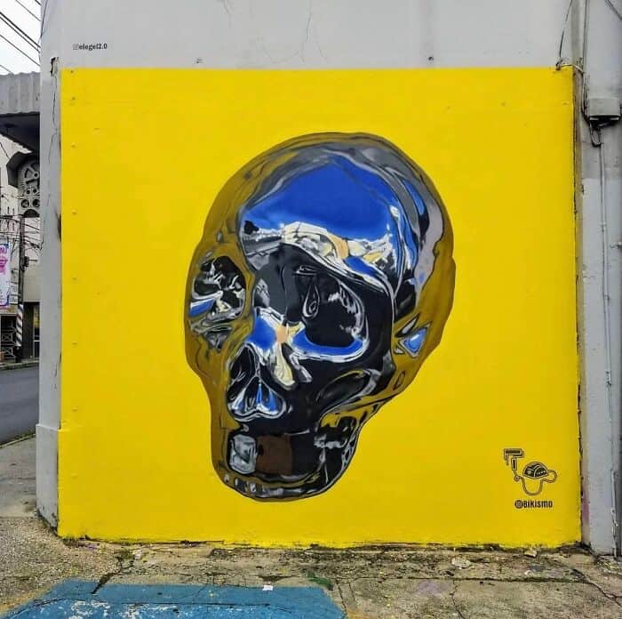 A vibrant yellow wall featuring a giant, hyper-realistic chrome skull. The artist, Bikismo, utilized expert shading to create a mirror-like effect, where the "metal" surface of the skull appears to reflect the entire street and the blue sky above, making the 2D surface look like a solid, polished object.