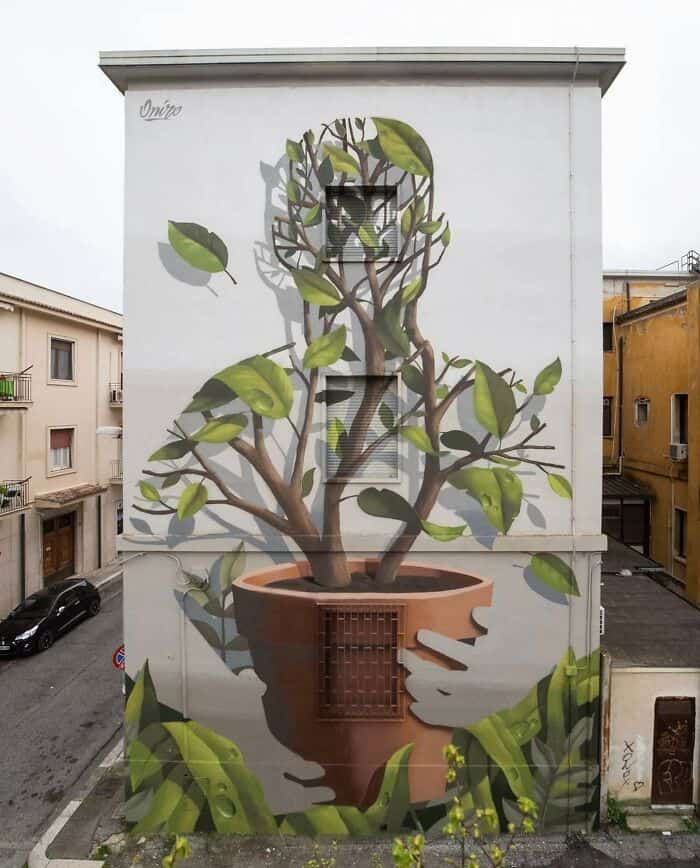 A poetic mural by Oniro that uses negative space and symbolism. A large terracotta pot is held by two ghostly grey hands; out of the pot grows a leafy tree whose branches and foliage are meticulously shaped to form the silhouette of a human head, representing the growth of consciousness.