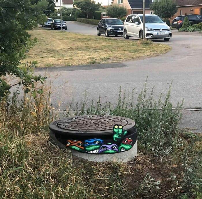 A clever example of urban intervention. The concrete base of a manhole cover features amazing street art in a pixelated style. The four Teenage Mutant Ninja Turtles—Leonardo, Raphael, Donatello, and Michelangelo—are painted peeking out from the "sewer," with Michelangelo throwing up a peace sign.