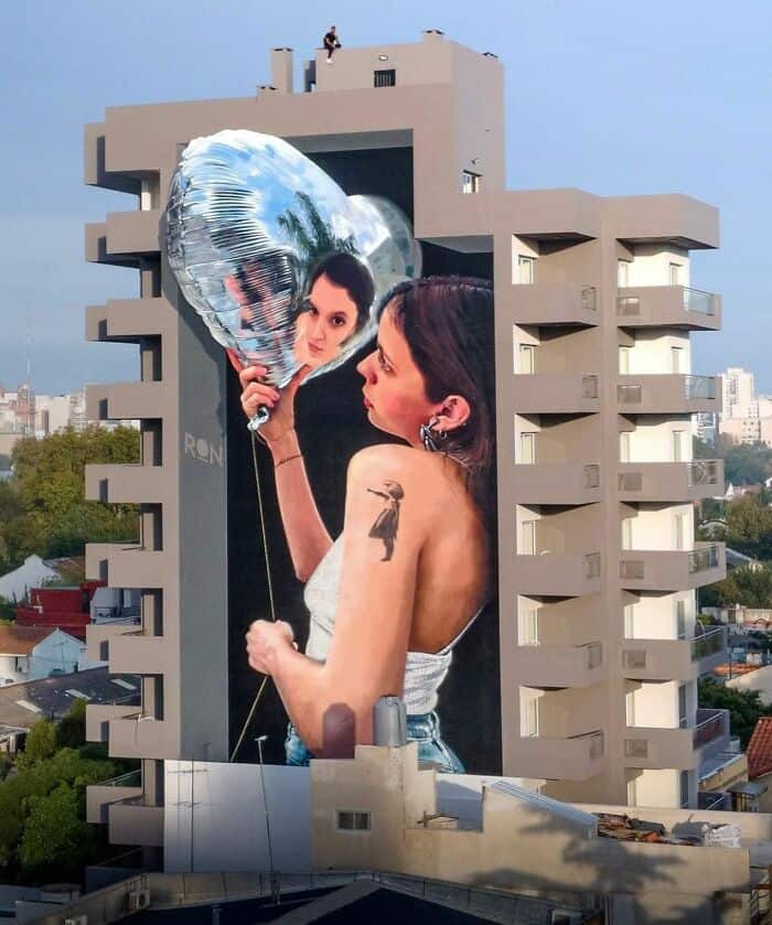 A breathtaking 3D mural on the side of a high-rise apartment building. The artwork depicts a woman holding a heart-shaped, reflective chrome balloon that mirrors her own face back at her. In a meta-tribute to street art history, she has a tattoo of Banksy’s iconic "Girl with Balloon" on her shoulder.