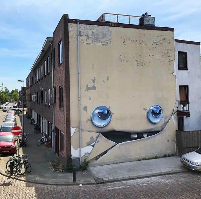 Creative pareidolia at its finest. This amazing street art transforms a dilapidated, peeling yellow wall into a giant, whimsical character. Large, realistic blue eyes are painted above a jagged horizontal crack in the stucco, which serves as a wide, toothy grin, turning urban decay into a playful face.