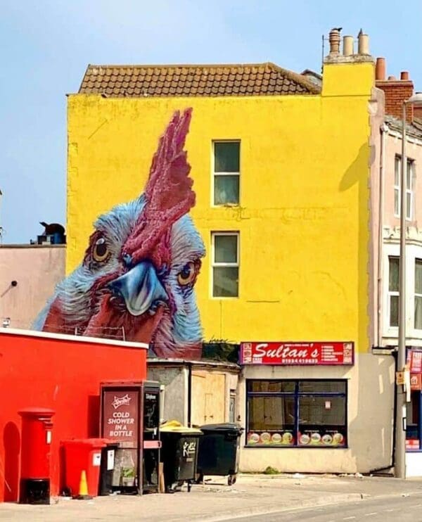 A bold and colorful mural of a giant rooster peeking over a building in an industrial area. The rooster’s detailed head and bright red comb contrast sharply against a bright yellow wall. The perspective makes it look as though the bird is curiously observing the street below near a shop called "Sultan's."