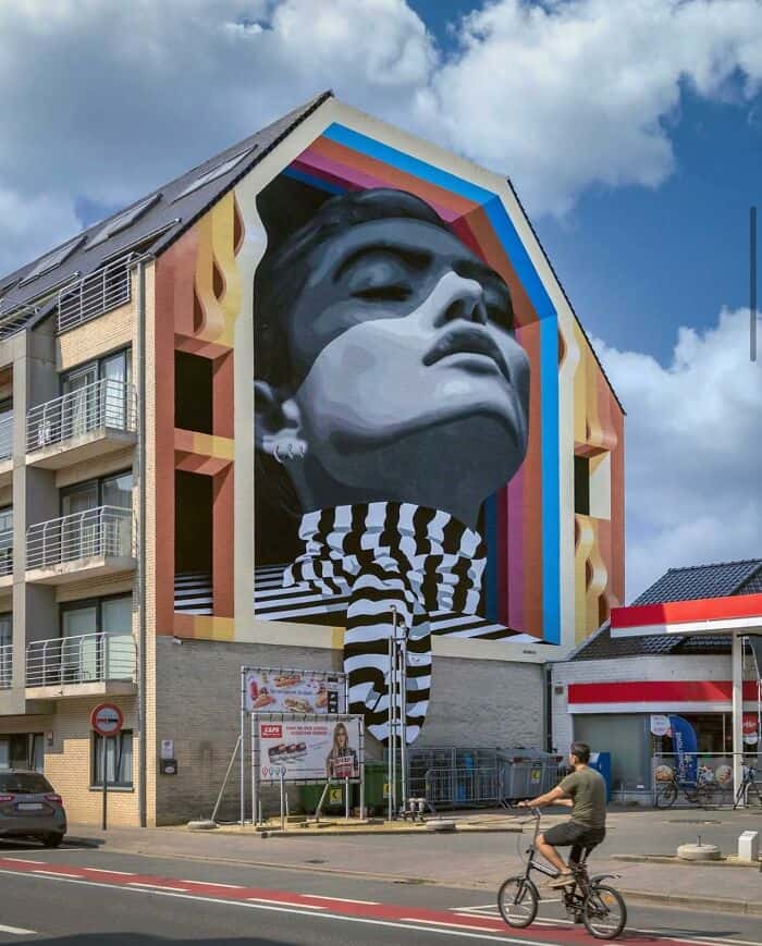 An incredible example of anamorphic amazing street art on an apartment building. A woman with her head tilted back is painted within a recessed "frame" that uses forced perspective to look like a room. Her black-and-white striped turtleneck appears to "spill" out of the mural and down the side of the building toward the sidewalk.