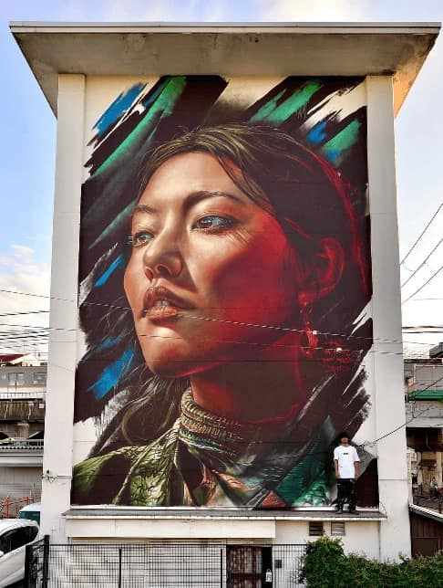 A massive, hyper-realistic portrait of a woman with a serene expression, towering over a city street. The mural features warm, sun-kissed lighting on her face and is framed by dynamic, abstract teal and black brushstrokes. A real person standing on the ledge at the bottom emphasizes the monumental scale of the piece.