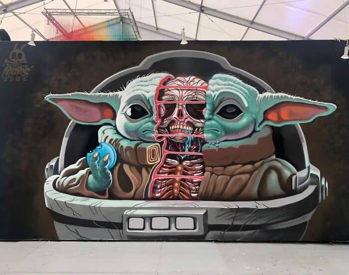 A surrealist take on pop culture. This amazing street art shows Grogu (Baby Yoda) sitting in his floating pod holding a blue cookie. However, the character is "split" open down the center to reveal a detailed, anatomical skeletal and muscular structure inside, blending fan art with lowbrow "dissected" aesthetics.