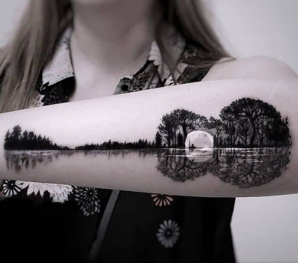 Forearm tattoo of a forest landscape and moon reflected in water, shaped like an acoustic guitar.