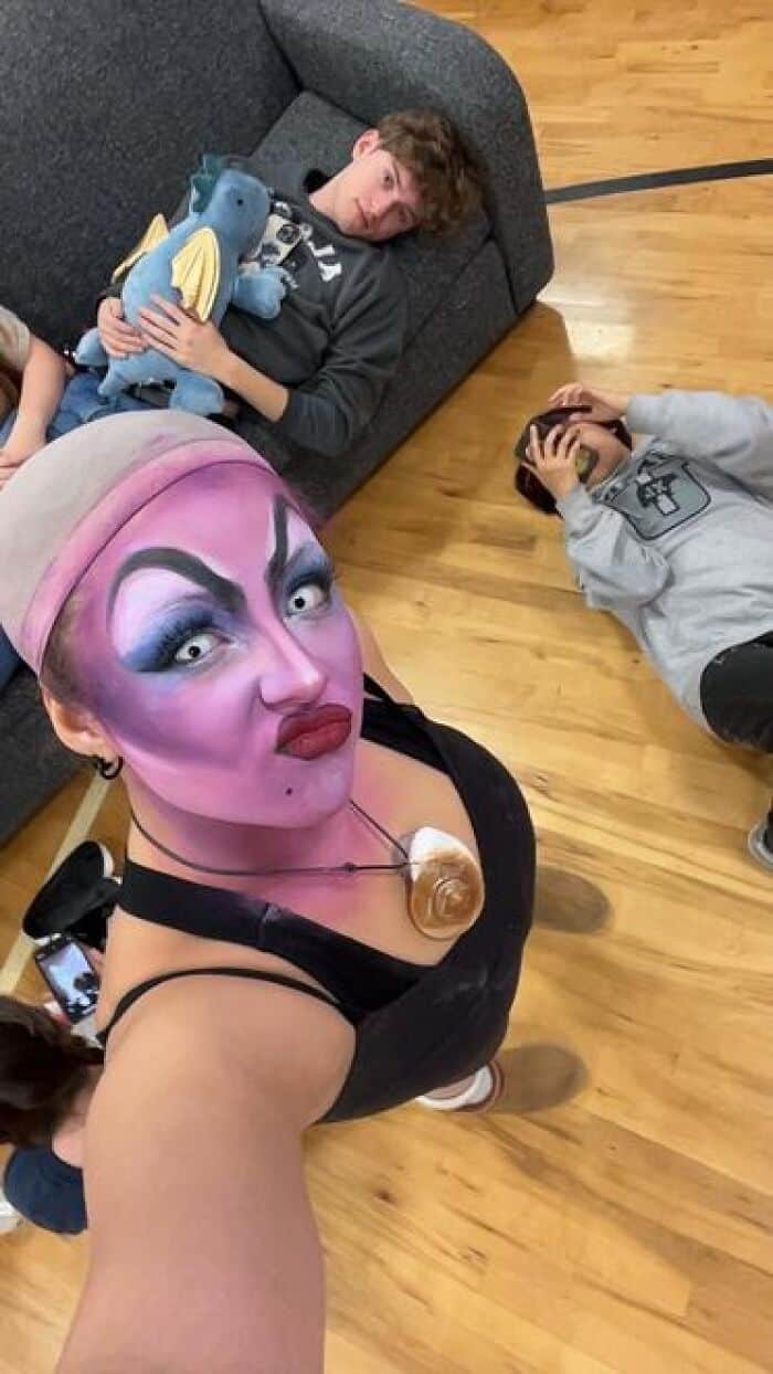 Student in bold Ursula-inspired purple drag stage makeup takes selfie while exhausted castmates rest on floor