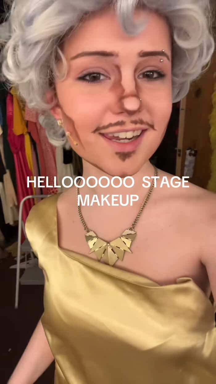 Young performer in gold dress and silver wig captioned "helloooooo stage makeup" showing heavy theatrical contouring