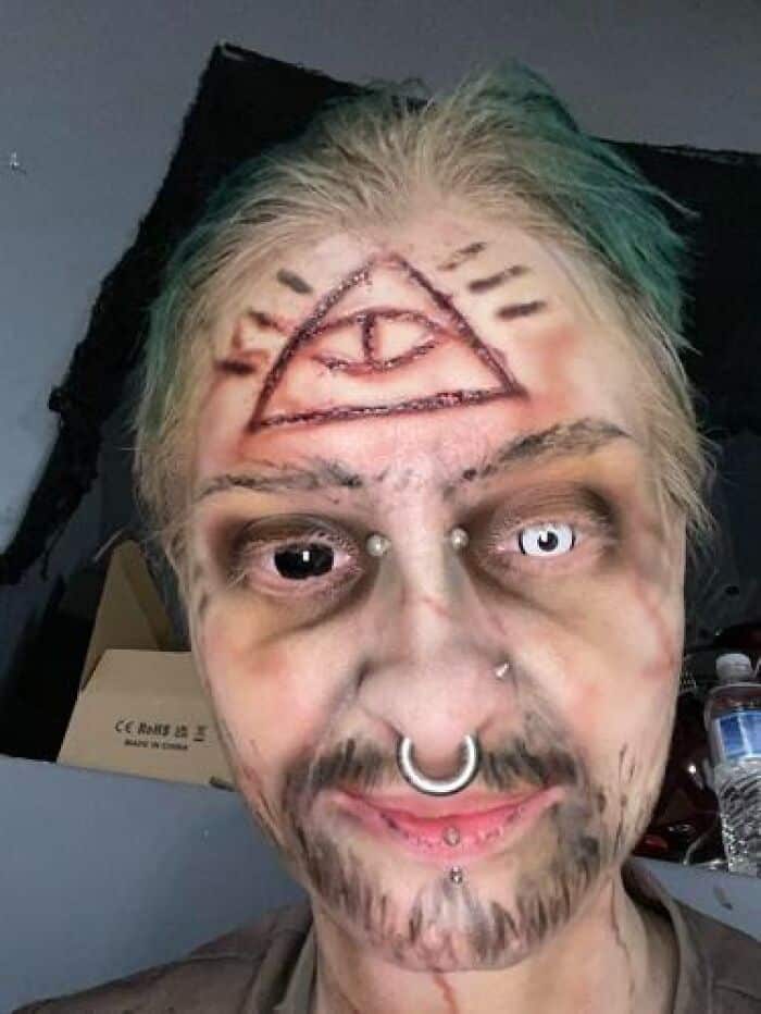 Performer wearing intense horror stage makeup featuring mismatched contact lenses and illuminati triangle forehead symbol