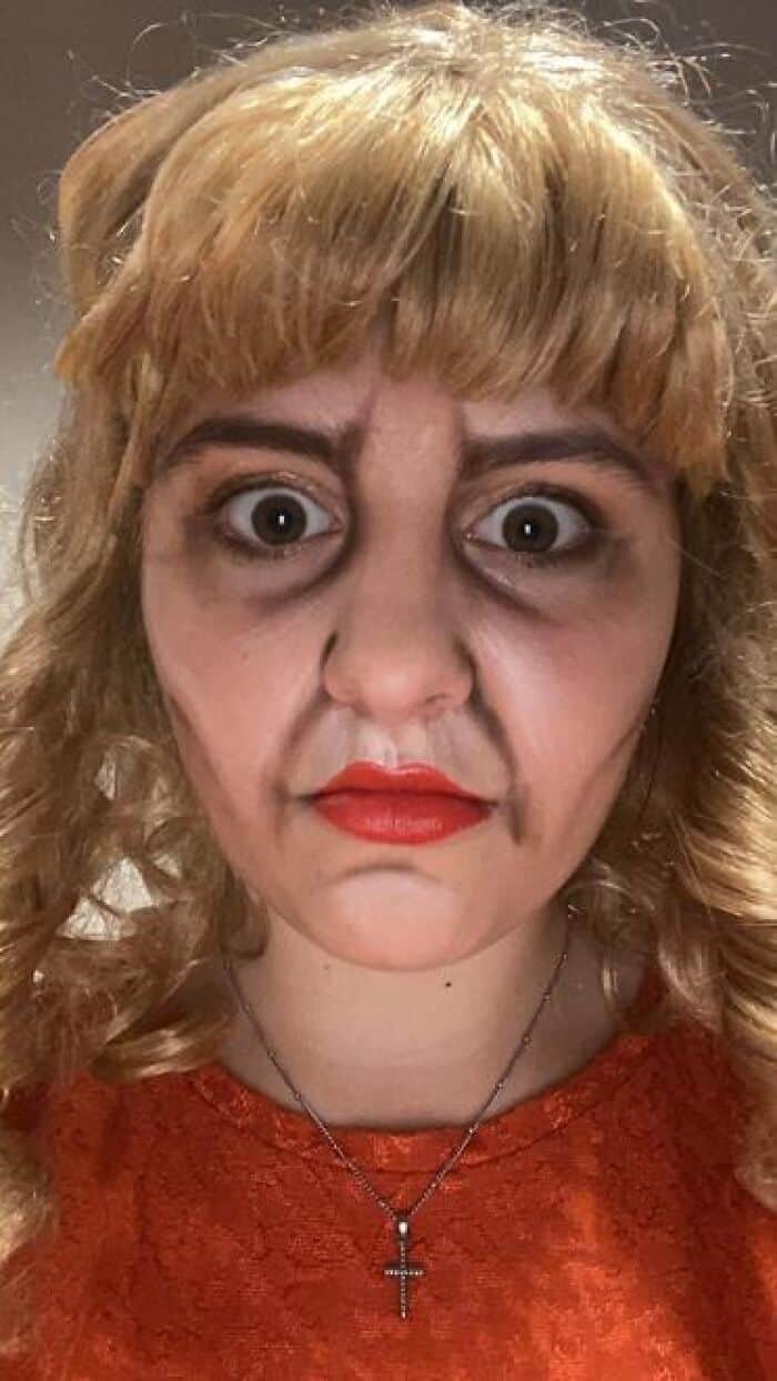 Performer in blonde wig wearing dramatic wide-eyed horror stage makeup with dark under-eye circles and red lips