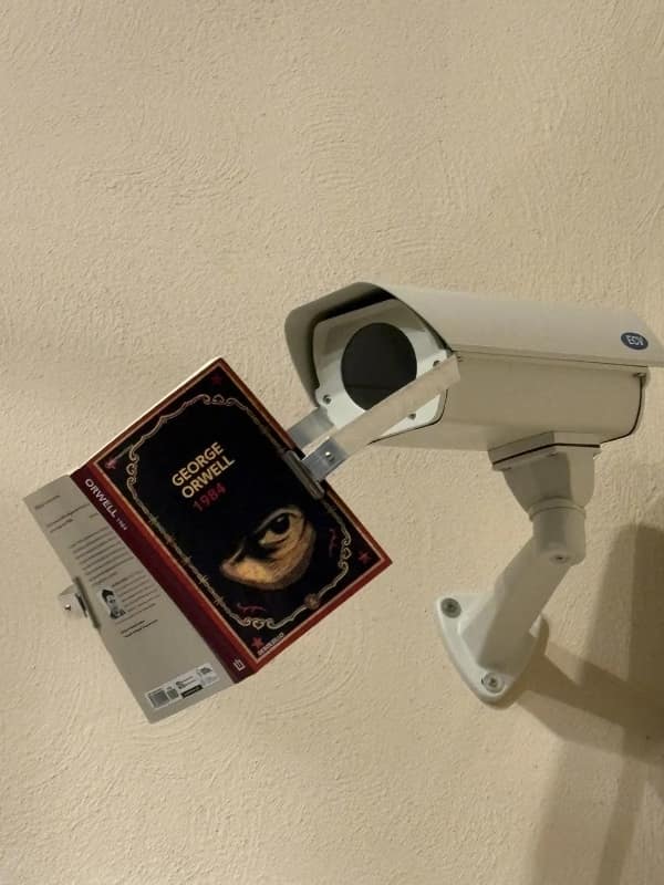 blursed image of a white industrial security camera that has been modified with a metal bracket to hold a copy of George Orwell’s 1984 directly in front of its lens. The camera appears to be intensely "studying" the foundational text of the surveillance state it serves.