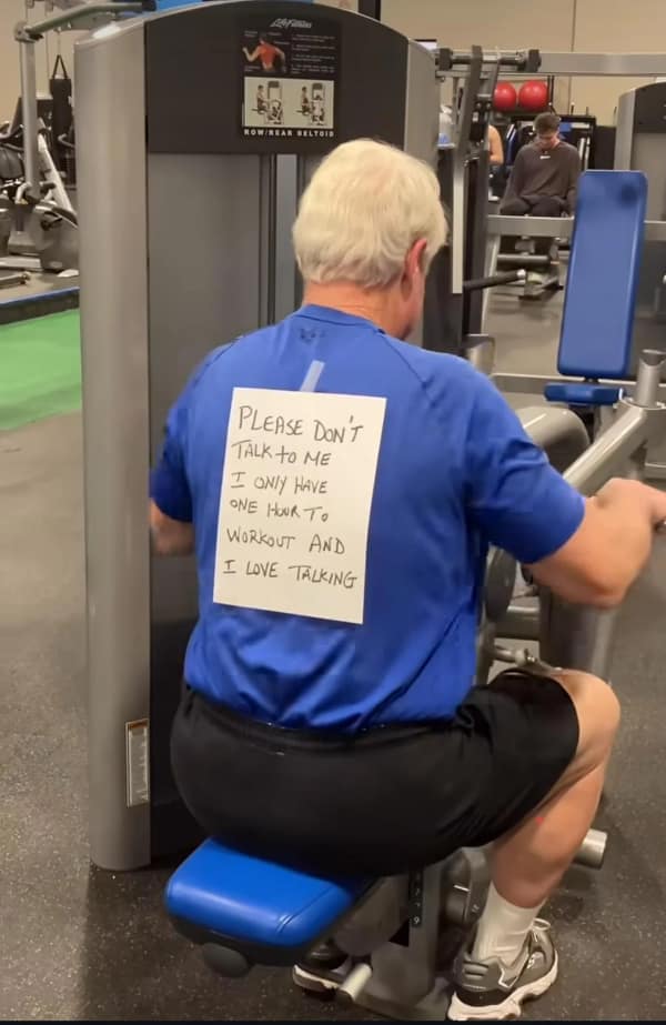 A relatable blursed image of an older man at a gym performing a rowing exercise. Taped to his blue t-shirt is a handwritten sign that reads, "PLEASE DON'T TALK TO ME I ONLY HAVE ONE HOUR TO WORKOUT AND I LOVE TALKING," capturing the struggle of a social butterfly trying to be productive.