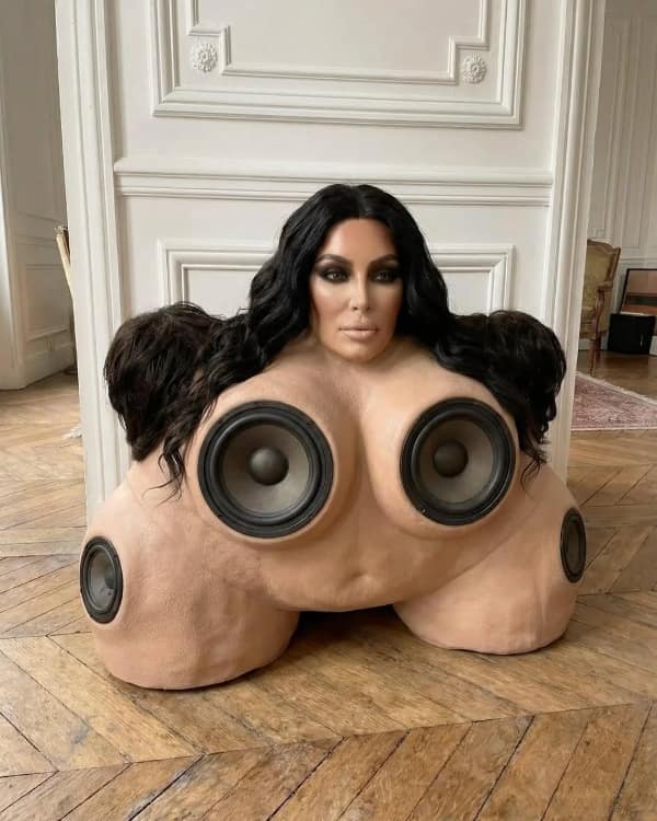 A nightmare-inducing blursed image of a surreal art installation. It features Kim Kardashian's face atop a fleshy, skin-toned structure embedded with multiple high-powered subwoofer speakers where limbs or breasts should be.