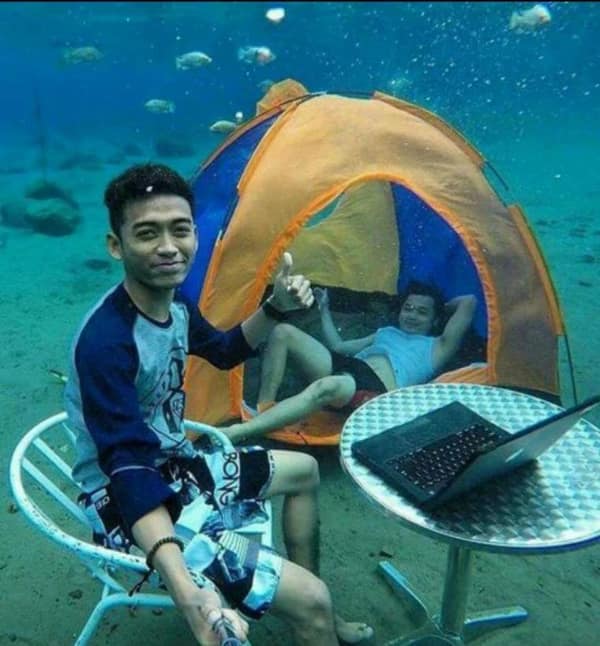 blursed image showing two men fully submerged at the bottom of a tropical ocean. They have a bright orange camping tent set up on the sand and a silver café table with a functioning laptop, posing with a thumbs-up as fish swim by.